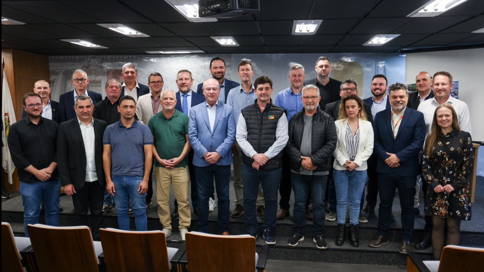 Grupo de aproximadamente 25 pessoas posa para foto em auditório, dispostas em três fileiras. A maioria veste roupas formais, e ao fundo há uma parede com imagem em tons de cinza.