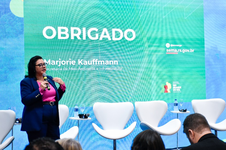 A imagem mostra a secretária em pé diante de um púlpito transparente, segurando um microfone. Ao fundo, a tela exibe a palavra “OBRIGADO” em letras grandes, seguida pelo nome “Marjorie Kauffmann” e a identificação “Secretaria de Meio Ambiente e Infraestrutura”, além do endereço eletrônico “sema.rs.gov.br” e o logotipo do Governo do Estado do Rio Grande do Sul.