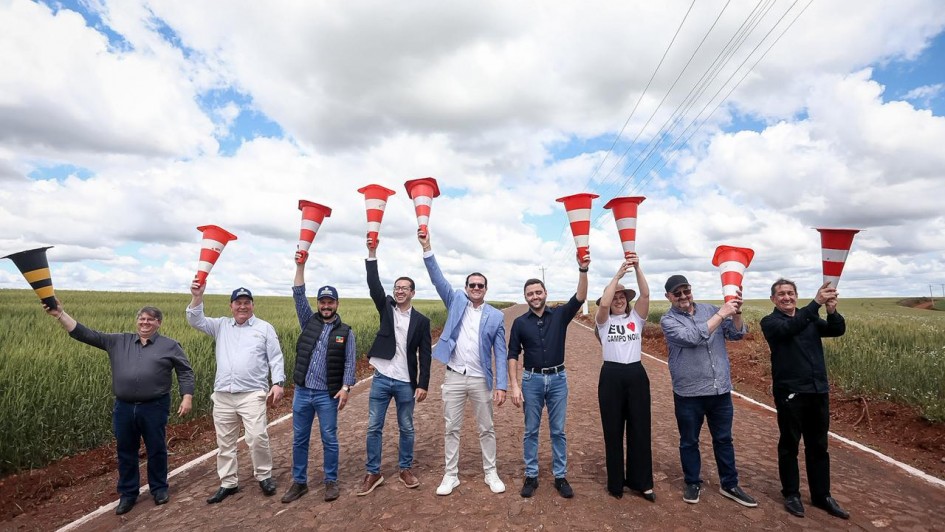 Um grupo de dez pessoas está posando em uma estrada rural pavimentada, sob um céu com nuvens e áreas de céu azul. Todas estão alinhadas e seguram cones de sinalização laranja e brancos erguidos acima da cabeça, em gesto de comemoração. Ao fundo, há plantações verdes dos dois lados da estrada. O clima é de celebração, e as pessoas sorriem, voltadas para a câmera.