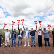 Um grupo de dez pessoas está posando em uma estrada rural pavimentada, sob um céu com nuvens e áreas de céu azul. Todas estão alinhadas e seguram cones de sinalização laranja e brancos erguidos acima da cabeça, em gesto de comemoração. Ao fundo, há plantações verdes dos dois lados da estrada. O clima é de celebração, e as pessoas sorriem, voltadas para a câmera.