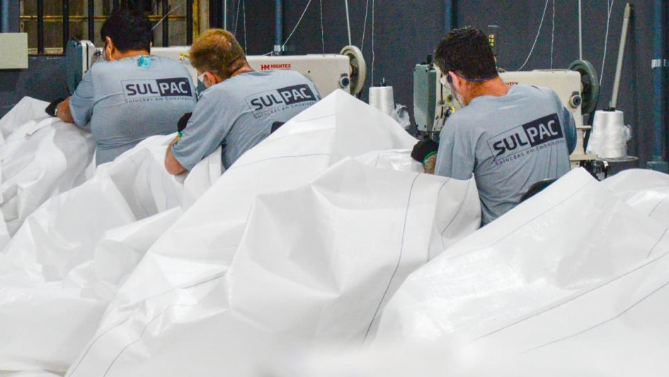 A imagem mostra três pessoas trabalhando em uma linha de produção têxtil, manipulando grandes peças de tecido branco em máquinas de costura industriais. Todas vestem camisetas cinza com o logotipo “SULPAC” estampado nas costas. O ambiente é interno, com paredes escuras e iluminação artificial.