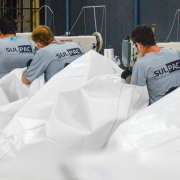 A imagem mostra três pessoas trabalhando em uma linha de produção têxtil, manipulando grandes peças de tecido branco em máquinas de costura industriais. Todas vestem camisetas cinza com o logotipo “SULPAC” estampado nas costas. O ambiente é interno, com paredes escuras e iluminação artificial.