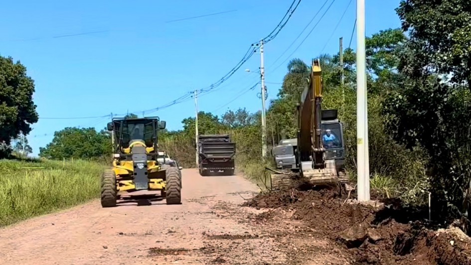 A imagem mostra uma estrada de terra sob céu limpo e azul, onde ocorre uma obra de infraestrutura. À direita, uma escavadeira está operando próxima a um poste de energia elétrica, removendo terra e vegetação da margem da estrada. Um trabalhador pode ser visto dentro da cabine da escavadeira. Há também um caminhão basculante mais ao fundo, aparentemente esperando para carregar ou descarregar materiais. À esquerda da estrada, um motoniveladora (patrol) amarela e preta avança no sentido da câmera, possivelmente nivelando o solo. A via está parcialmente coberta por terra solta, sugerindo que os trabalhos estão em andamento para melhorar ou alargar a estrada. Vegetação verde margeia ambos os lados da via.