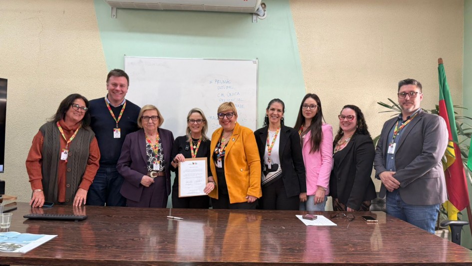 Nove pessoas estão alinhadas atrás de uma mesa de madeira em uma sala de reuniões. O grupo, formado por homens e mulheres, sorri em direção à câmera. No centro, duas mulheres seguram um documento. Ao fundo, há um quadro branco com anotações e, à direita, parte de uma bandeira do Rio Grande do Sul. O ambiente tem iluminação interna e aparência institucional.