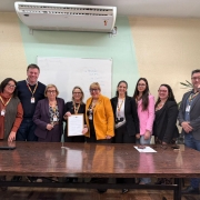 Nove pessoas estão alinhadas atrás de uma mesa de madeira em uma sala de reuniões. O grupo, formado por homens e mulheres, sorri em direção à câmera. No centro, duas mulheres seguram um documento. Ao fundo, há um quadro branco com anotações e, à direita, parte de uma bandeira do Rio Grande do Sul. O ambiente tem iluminação interna e aparência institucional.
