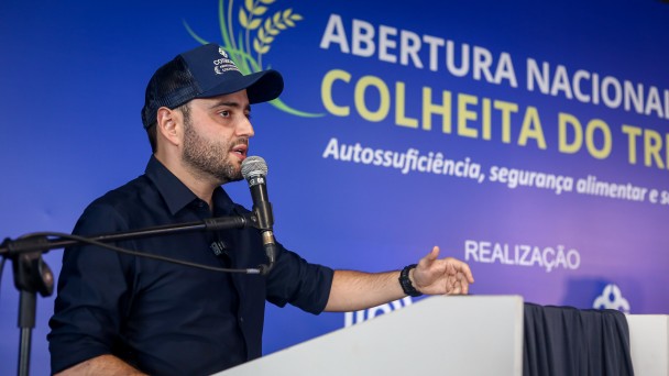 O vice-governador Gabriel Souza aparece discursando em um púlpito, usando boné azul e camisa escura. Ao fundo, painel azul com os dizeres “Abertura Nacional da Colheita do Trigo”.