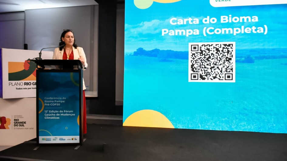 Mulher fala ao microfone em um púlpito sobre um palco, com fundo iluminado por um grande painel digital azul. O painel exibe o texto “Carta do Bioma Pampa (Completa)” e um código QR. Ao lado do púlpito há um banner do “Plano Rio Grande” com o logotipo do Governo do Estado do Rio Grande do Sul.