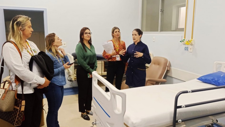 Cinco mulheres estão em uma enfermaria hospitalar, ao lado de um leito com lençol branco. Uma profissional de jaleco azul fala e gesticula, enquanto as demais ouvem atentamente, algumas com documentos e celulares nas mãos.