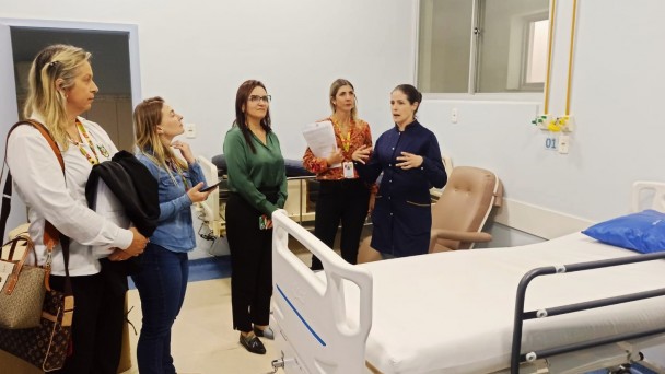 Cinco mulheres estão em uma enfermaria hospitalar, ao lado de um leito com lençol branco. Uma profissional de jaleco azul fala e gesticula, enquanto as demais ouvem atentamente, algumas com documentos e celulares nas mãos.