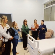 Cinco mulheres estão em uma enfermaria hospitalar, ao lado de um leito com lençol branco. Uma profissional de jaleco azul fala e gesticula, enquanto as demais ouvem atentamente, algumas com documentos e celulares nas mãos.