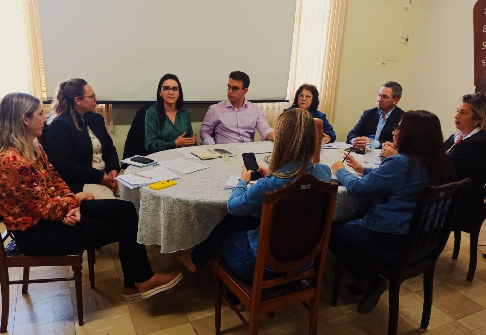 Grupo de nove pessoas participa de uma reunião em torno de uma mesa redonda coberta por toalha branca. Há cadernos, celulares e documentos sobre a mesa. Uma mulher de blusa verde fala enquanto os demais acompanham a discussão.