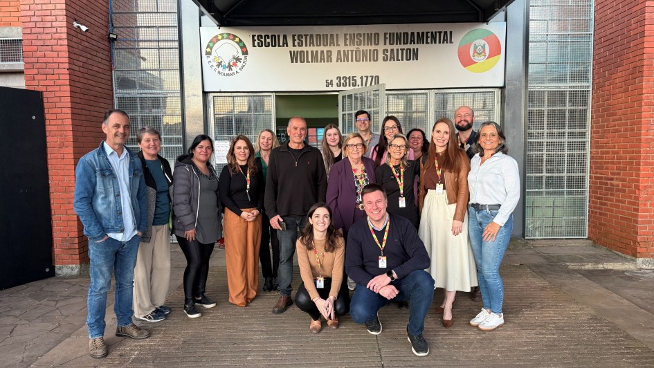 otografia de um grupo composto por dezesseis pessoas posando em frente à entrada principal de um prédio escolar. Acima da porta, há uma placa com os dizeres "ESCOLA ESTADUAL ENSINO FUNDAMENTAL WOLMAR ANTÔNIO SALTON" e o número de telefone "54 3315.1771". A placa também exibe dois logotipos, um à esquerda e outro à direita. O grupo está organizado em duas fileiras: algumas pessoas estão agachadas na frente, enquanto outras permanecem em pé atrás. O cenário indica que se trata da fachada principal da escola mencionada.
