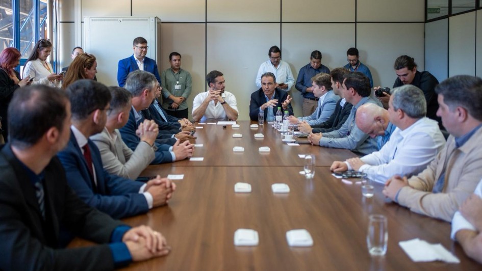 Reunião com autoridades e técnicos em uma sala de reuniões. O governador Eduardo Leite está ao centro, vestindo camisa branca, gesticulando enquanto conversa com o grupo. À sua direita, outro participante fala enquanto os demais ouvem atentos.