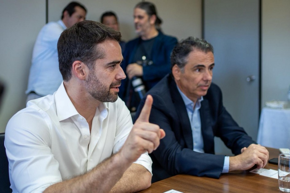 O governador Eduardo Leite aparece em primeiro plano, à esquerda, vestindo camisa branca e falando com ênfase, com um gesto de mão levantada. Ao seu lado direito, outro homem de terno azul o acompanha atentamente. Ao fundo, pessoas observam e registram o momento.
