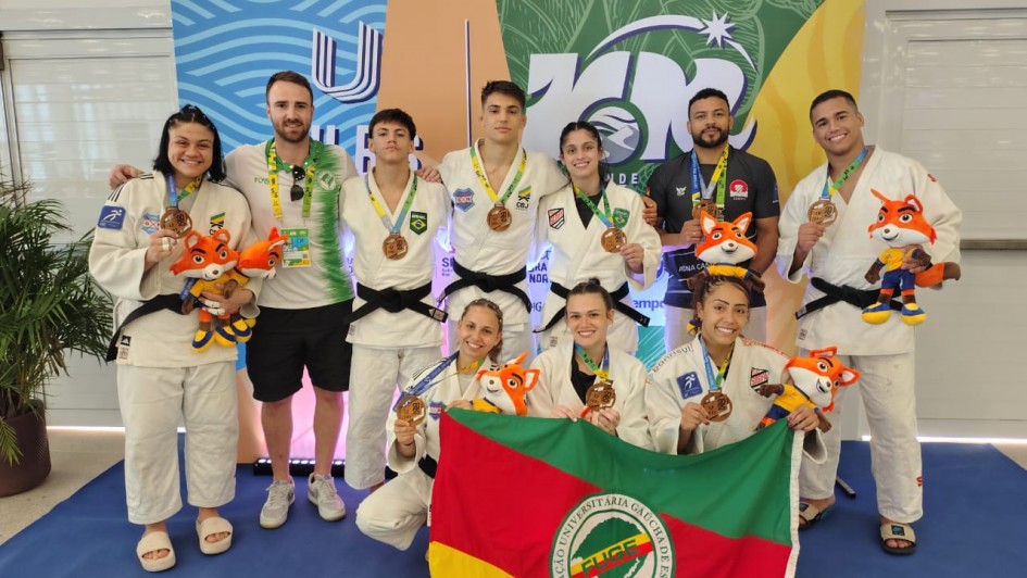 Um grupo de dez pessoas posa para uma foto em frente a um banner colorido, com os dizeres "JUBs" (Jogos Universitários Brasileiros) e o número "100" estilizado, indicando o centenário da competição. O grupo é composto por atletas de judô, vestidos com quimonos brancos (judogis), e alguns técnicos ou membros da equipe de apoio.
A maioria dos atletas exibe medalhas no pescoço e segura mascotes de pelúcia, representando um personagem laranja com roupas esportivas, provavelmente o mascote oficial do evento. Três atletas estão ajoelhados na frente, enquanto os demais estão de pé atrás deles. Duas atletas ajoelhadas seguram uma bandeira verde, vermelha e amarela com o logotipo da FUGE (Federação Universitária Gaúcha de Esportes), indicando que representam o estado do Rio Grande do Sul.
Todos na imagem parecem felizes e orgulhosos, comemorando suas conquistas no evento esportivo.