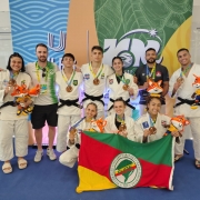 Um grupo de dez pessoas posa para uma foto em frente a um banner colorido, com os dizeres "JUBs" (Jogos Universitários Brasileiros) e o número "100" estilizado, indicando o centenário da competição. O grupo é composto por atletas de judô, vestidos com quimonos brancos (judogis), e alguns técnicos ou membros da equipe de apoio.

A maioria dos atletas exibe medalhas no pescoço e segura mascotes de pelúcia, representando um personagem laranja com roupas esportivas, provavelmente o mascote oficial do evento. Três atletas estão ajoelhados na frente, enquanto os demais estão de pé atrás deles. Duas atletas ajoelhadas seguram uma bandeira verde, vermelha e amarela com o logotipo da FUGE (Federação Universitária Gaúcha de Esportes), indicando que representam o estado do Rio Grande do Sul.

Todos na imagem parecem felizes e orgulhosos, comemorando suas conquistas no evento esportivo.