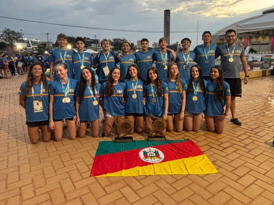 A imagem mostra um grupo de 22 jovens atletas (homens e mulheres) posando para uma foto coletiva ao ar livre, sobre um chão de tijolos aparentes. Eles vestem camisetas azuis com detalhes em laranja e amarelo, com a sigla "JEBs" (Jogos Escolares Brasileiros) estampada. A maioria deles está com medalhas douradas e prateadas penduradas no pescoço, indicando que participaram de competições esportivas e conquistaram prêmios.
Na frente do grupo, no chão, estão expostos dois troféus e uma bandeira do estado do Rio Grande do Sul, com as cores verde, vermelha e amarela, e o brasão no centro. A equipe é composta majoritariamente por adolescentes e jovens, acompanhados por um treinador ou técnico posicionado à direita.
Ao fundo, é possível ver outras pessoas, barracas e estruturas de um evento esportivo, com iluminação artificial acesa ao anoitecer. O clima da imagem é de celebração, conquista e união.