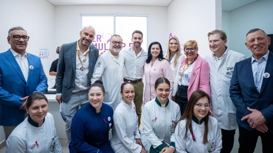 A foto mostra um grupo de pessoas reunidas em um ambiente interno de hospital. Ao fundo, há uma janela com adesivo escrito “SER MULHER RS” em letras roxas. As pessoas estão vestindo roupas formais e jalecos. O governador está ao centro do grupo.