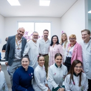 A foto mostra um grupo de pessoas reunidas em um ambiente interno de hospital. Ao fundo, há uma janela com adesivo escrito “SER MULHER RS” em letras roxas. As pessoas estão vestindo roupas formais e jalecos. O governador está ao centro do grupo. 