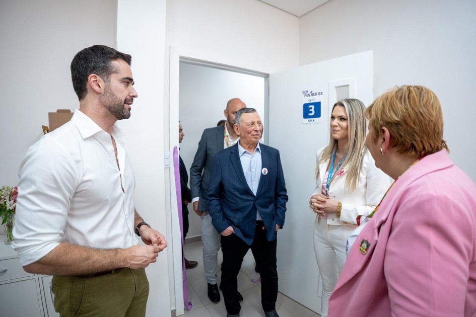 A imagem retrata um grupo de pessoas conversando com o governador em uma sala interna, com paredes brancas e portas identificadas por placas azuis. Uma das portas tem a inscrição “Sala 3” e o logotipo “SER MULHER RS”.