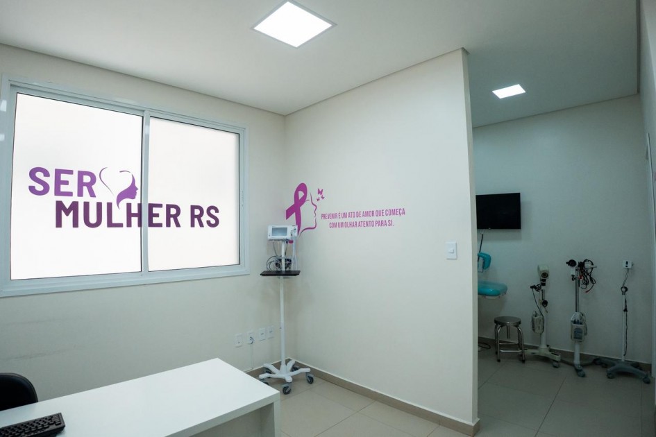 A foto mostra uma sala clara e organizada, com paredes brancas e piso bege. À esquerda, há uma janela com adesivo grande escrito “SER MULHER RS” em roxo. Na parede lateral, há uma frase motivacional acompanhada do laço rosa, símbolo da prevenção ao câncer de mama. A sala contém uma mesa branca, um suporte com equipamento médico e, ao fundo, uma área com aparelhos hospitalares.