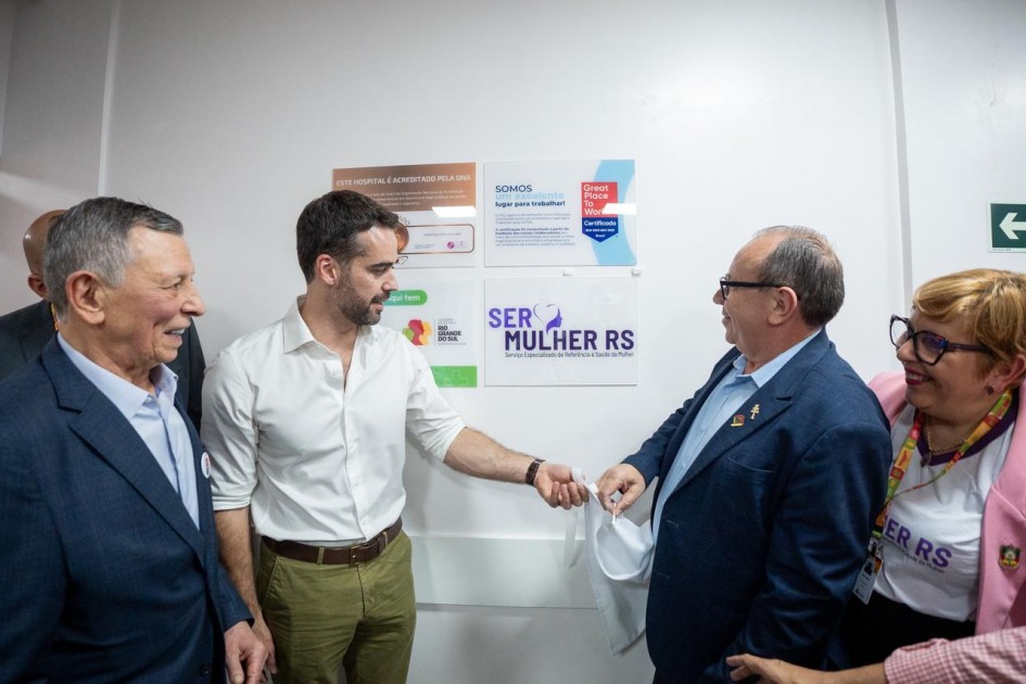 A imagem apresenta um grupo de pessoas em pé diante de uma parede branca com cartazes fixados, com o governador ao centro. Um dos cartazes traz o texto “SER MULHER RS” em destaque, junto a um símbolo roxo. Outros cartazes exibem informações sobre serviços e campanhas.