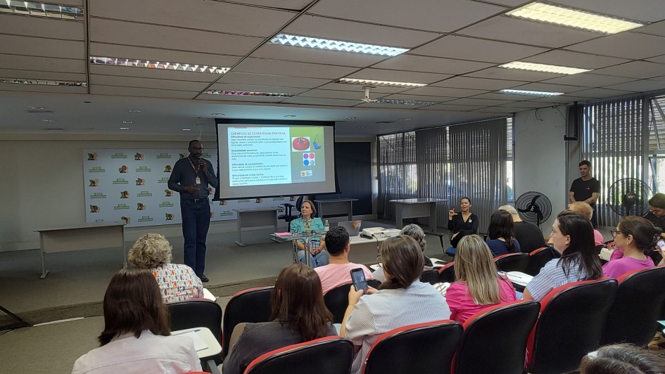 A imagem mostra um palestrante em pé em frente a um auditório com diversas pessoas sentadas, assistindo a apresentação. Ao fundo, há uma tela com a apresentação em andamento.
