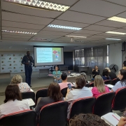 A imagem mostra um palestrante em pé em frente a um auditório com diversas pessoas sentadas, assistindo a apresentação. Ao fundo, há uma tela com a apresentação em andamento.