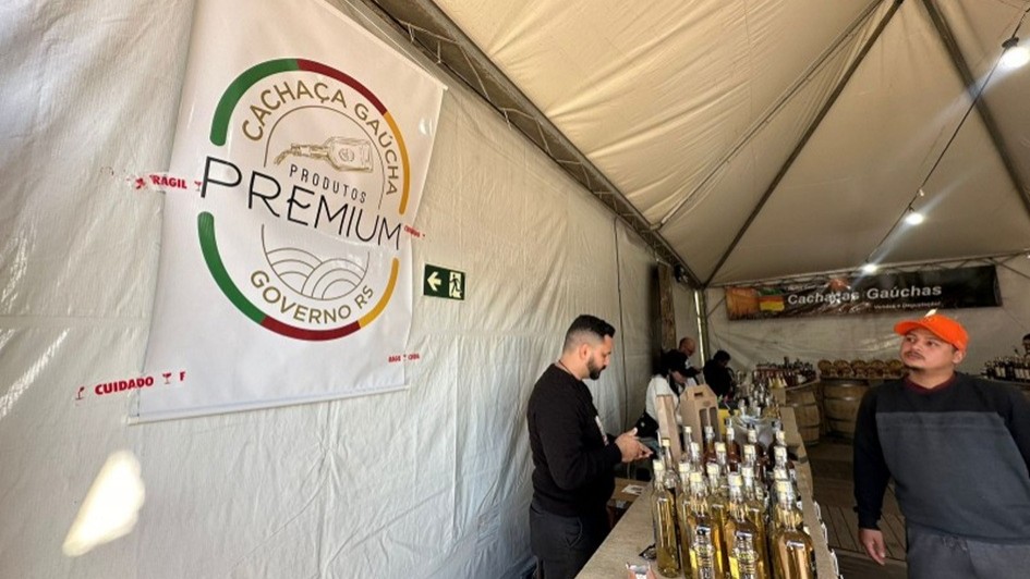 A imagem mostra o interior de uma tenda de feira ou evento, dedicada &agrave; exposi&ccedil;&atilde;o e venda de cacha&ccedil;as ga&uacute;chas. &Agrave; esquerda, h&aacute; um grande banner branco com o logotipo "Cacha&ccedil;a Ga&uacute;cha - Produtos Premium - Governo RS", destacando que s&atilde;o produtos selecionados e de qualidade, promovidos pelo governo do Rio Grande do Sul.

No centro da imagem, h&aacute; uma mesa comprida coberta com tecido r&uacute;stico (parecido com juta), onde est&atilde;o dispostas diversas garrafas de cacha&ccedil;a, em diferentes tamanhos e r&oacute;tulos. Tamb&eacute;m h&aacute; um pequeno item decorativo em forma de bota ga&uacute;cha em cima da mesa.

Atr&aacute;s da mesa, est&atilde;o algumas pessoas, incluindo um homem de camisa escura manuseando algo e outro homem com bon&eacute; laranja e moletom cinza escuro. Ao fundo, outro banner preto com a inscri&ccedil;&atilde;o "Cacha&ccedil;as Ga&uacute;chas" pode ser visto acima de mais barris e garrafas expostas.

O ambiente sugere um espa&ccedil;o de degusta&ccedil;&atilde;o e venda de cacha&ccedil;as artesanais em um evento regional, provavelmente voltado &agrave; valoriza&ccedil;&atilde;o de produtos locais.