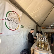 A imagem mostra o interior de uma tenda de feira ou evento, dedicada à exposição e venda de cachaças gaúchas. À esquerda, há um grande banner branco com o logotipo "Cachaça Gaúcha - Produtos Premium - Governo RS", destacando que são produtos selecionados e de qualidade, promovidos pelo governo do Rio Grande do Sul.

No centro da imagem, há uma mesa comprida coberta com tecido rústico (parecido com juta), onde estão dispostas diversas garrafas de cachaça, em diferentes tamanhos e rótulos. Também há um pequeno item decorativo em forma de bota gaúcha em cima da mesa.

Atrás da mesa, estão algumas pessoas, incluindo um homem de camisa escura manuseando algo e outro homem com boné laranja e moletom cinza escuro. Ao fundo, outro banner preto com a inscrição "Cachaças Gaúchas" pode ser visto acima de mais barris e garrafas expostas.

O ambiente sugere um espaço de degustação e venda de cachaças artesanais em um evento regional, provavelmente voltado à valorização de produtos locais.