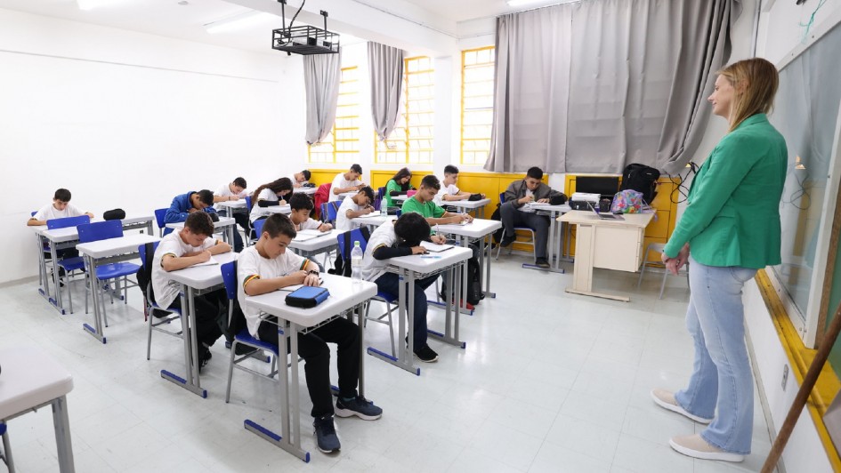 Professora em pé em sala de aula, para alunos que acompanham sentados em suas classes. 