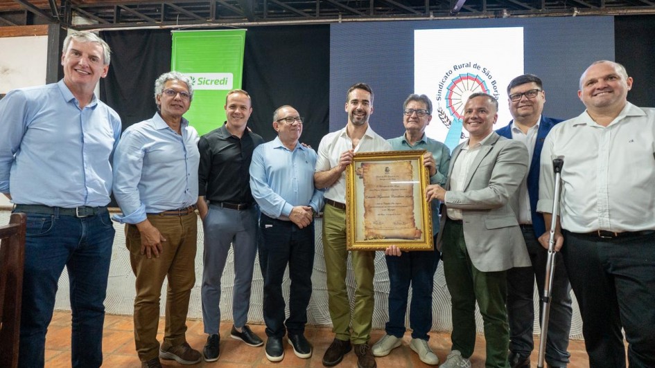 Um grupo de homens está reunido em pé, posando para a foto. Ao centro, segurando um quadro com moldura dourada, está o governador.