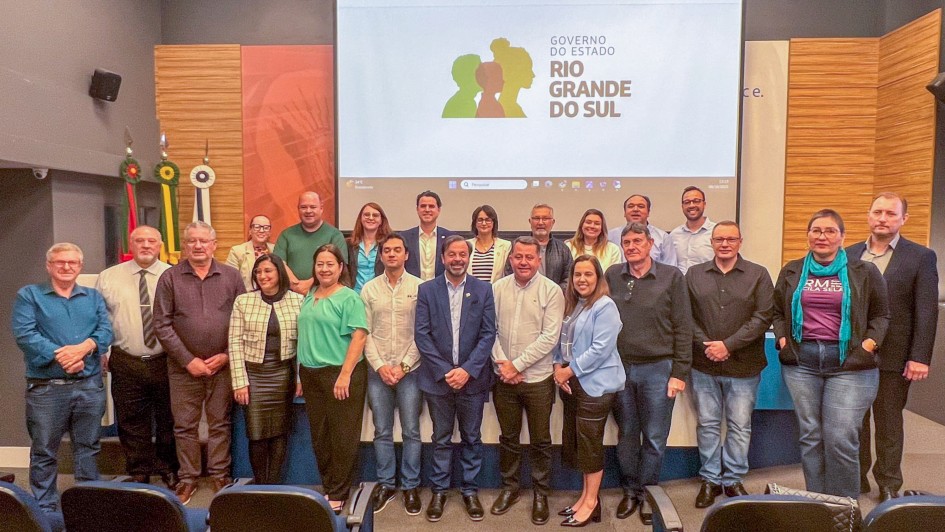 Vinte e uma pessoas estão reunidas em duas fileiras, posando para uma foto em ambiente interno, o auditório da Federação das Associações de Municípios do Rio Grande do Sul (Famurs). Ao fundo, há uma tela grande com o logotipo e o texto “Governo do Estado Rio Grande do Sul”. O logotipo mostra três silhuetas de perfis humanos nas cores verde, amarela e vermelha. À esquerda da imagem, há bandeiras posicionadas verticalmente. As pessoas estão vestidas com roupas sociais ou esporte fino.