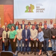 Vinte e uma pessoas estão reunidas em duas fileiras, posando para uma foto em ambiente interno, o auditório da Federação das Associações de Municípios do Rio Grande do Sul (Famurs). Ao fundo, há uma tela grande com o logotipo e o texto “Governo do Estado Rio Grande do Sul”. O logotipo mostra três silhuetas de perfis humanos nas cores verde, amarela e vermelha. À esquerda da imagem, há bandeiras posicionadas verticalmente. As pessoas estão vestidas com roupas sociais ou esporte fino.