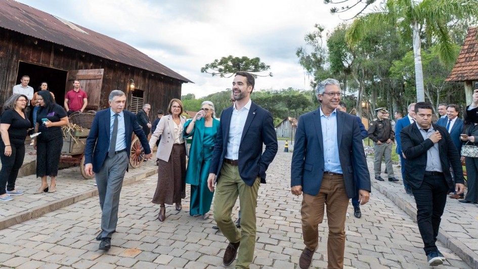 A imagem retrata um grupo caminhando sobre um caminho de pedras em um espaço aberto com construções de madeira ao redor. O governador está ao centro da imagem. Algumas pessoas estão vestidas com roupas sociais, enquanto outras usam trajes mais casuais. Ao fundo, há árvores altas, incluindo araucárias.