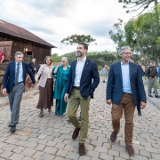 A imagem retrata um grupo caminhando sobre um caminho de pedras em um espaço aberto com construções de madeira ao redor. O governador está ao centro da imagem. Algumas pessoas estão vestidas com roupas sociais, enquanto outras usam trajes mais casuais. Ao fundo, há árvores altas, incluindo araucárias.