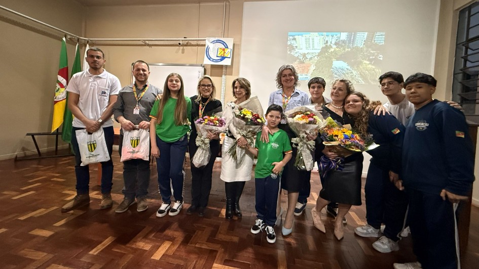 Grupo de pessoas reunidas em uma sala, com adultos e crianças sorrindo. Algumas certamente flores , há bandeiras ao fundo e uma tela com imagem de cidade. Parece uma cerimônia de homenagem ou celebração.