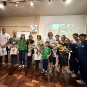 Grupo de pessoas reunidas em uma sala, com adultos e crianças sorrindo. Algumas certamente flores , há bandeiras ao fundo e uma tela com imagem de cidade. Parece uma cerimônia de homenagem ou celebração.