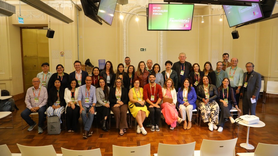 Grupo de cerca de 30 pessoas em sala elegante, com cadeiras brancas e telas exibindo temas de educação fiscal. Estão organizados para foto, muitos com crachás, diminuindo eventos profissionais ou educacionais.