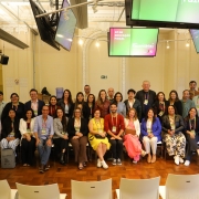 Grupo de cerca de 30 pessoas em sala elegante, com cadeiras brancas e telas exibindo temas de educação fiscal. Estão organizados para foto, muitos com crachás, diminuindo eventos profissionais ou educacionais.