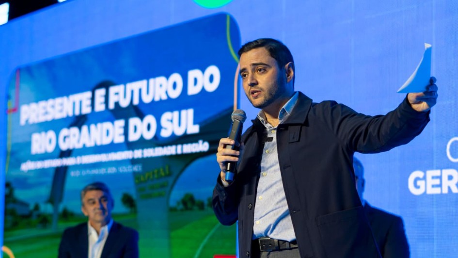 Homem em palco fala segurando papel, com tela ao fundo exibindo tema sobre saúde e desenvolvimento no Rio Grande do Sul. Outro homem está sentado, ambos em traje formal. Evento parece governamental.