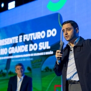 Homem em palco fala segurando papel, com tela ao fundo exibindo tema sobre saúde e desenvolvimento no Rio Grande do Sul. Outro homem está sentado, ambos em traje formal. Evento parece governamental.