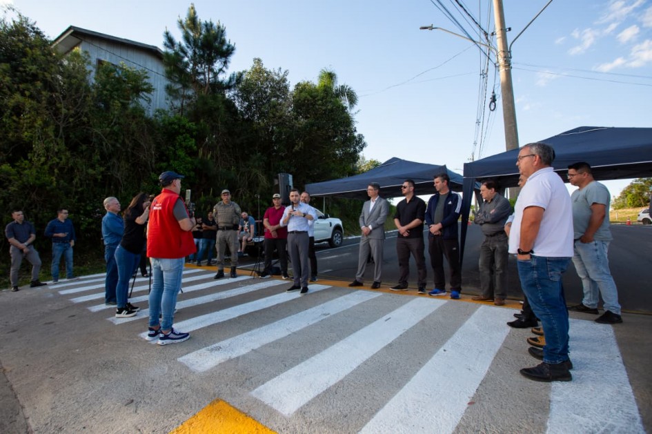 Grupo de pessoas reunidas em faixa de pedestres recém-pintada, ao lado das tendas azuis e carro branco. Evento ao ar livre, com falas e registros em vídeo, estreia ou ação comunitária.