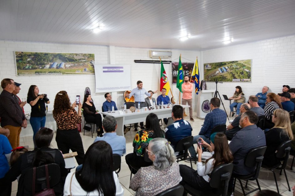 Reunião formal em sala clara, com autoridades sentadas à mesa e público atento. Há bandeiras, projeção com agenda do evento e fotos aéreas da área rural nas paredes.