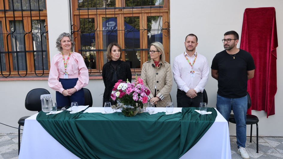 Cinco pessoas em pé atrás de mesa decorada com flores, em evento formal ao ar livre. Há microfones, copos e cortina vermelha cobrindo algo na parede, fazendo cerimônia ou anúncio público.