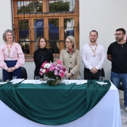 Cinco pessoas em pé atrás de mesa decorada com flores, em evento formal ao ar livre. Há microfones, copos e cortina vermelha cobrindo algo na parede, fazendo cerimônia ou anúncio público.