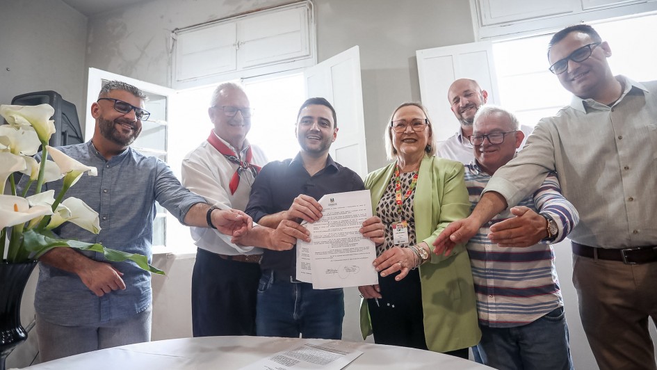 Seis pessoas sorridentes em pé ao redor de uma mesa com flores, celebrando a assinatura de um documento oficial. Ambiente claro e acolhedor, transmitindo união e conquista. 