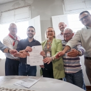 Seis pessoas sorridentes em pé ao redor de uma mesa com flores, celebrando a assinatura de um documento oficial. Ambiente claro e acolhedor, transmitindo união e conquista. 