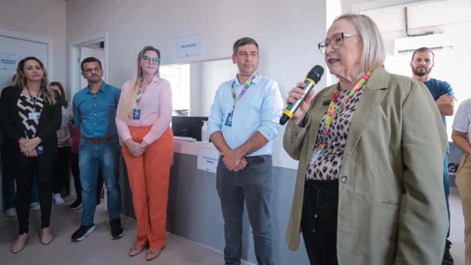 A imagem mostra um grupo de pessoas em um ambiente interno, provavelmente durante a inauguração de uma unidade de saúde pública. Em primeiro plano, à direita, uma mulher idosa de cabelos grisalhos usa óculos, um blazer verde claro e segura um microfone, aparentando estar discursando. Ao fundo, outras pessoas, homens e mulheres, estão em pé, ouvindo atentamente. Elas usam crachás, indicando que podem ser funcionários ou autoridades. Atrás deles, há um balcão com a placa "Recepção" e outra sinalização escrita "Atendimento Geral". O ambiente é claro e limpo, com paredes brancas e piso claro, sugerindo uma instalação recém-inaugurada ou reformada.