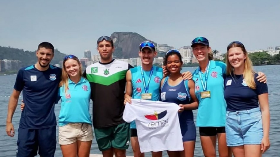 A imagem mostra sete pessoas em pé, ao ar livre, próximas a um corpo d'água, com prédios e uma montanha ao fundo. Elas estão vestidas com roupas esportivas e algumas usam medalhas no pescoço, indicando participação em um evento esportivo. Uma das pessoas segura uma camiseta branca com a palavra "remar" e a imagem de um remo. O ambiente está ensolarado, sugerindo que a foto foi tirada durante o dia.
